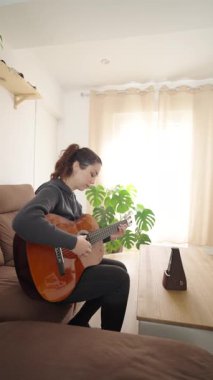 Kadın ritim için metronomu kullanarak gitar çalmaya odaklanıyor. Arka planda tropikal bir bitki bulunan rahat, iyi ışıklandırılmış bir oturma odasında oturuyor.