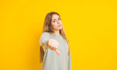 Studio shot of serious young woman showing thumbs down sign of disapproval, dislike or failure, expressing negative emotions against yellow background clipart