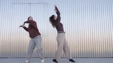 İki genç kadın, muhtemelen profesyonel dansçılar, kıvrımlı metal bir duvara karşı senkronize bir dans gösterisi yapıyorlar.