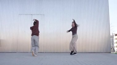 İki genç kadın, muhtemelen profesyonel dansçılar ya da öğrenciler, metalik bir duvarın önünde modern bir dans koreografisi yapıyorlar.