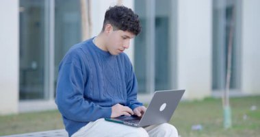 Kazaklı genç bir erkek öğrencinin gün ışığında kampüs binalarının yakınında otururken bilgisayarın ekranına bakması ve klavyeyle yazması gerçek zamanlı.