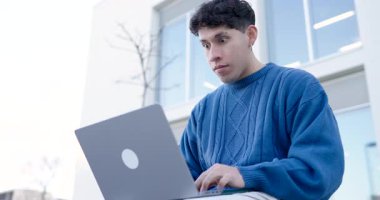 Gündüz vakti inşaata karşı otururken ani bir kayıp sonucu dizüstü bilgisayarda çalışan ciddi Latin kökenli serbest çalışan genç bir erkek.