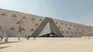 Büyük Mısır Müzesi 'nin ana girişinde gezen çeşitli turistler, giza kentindeki güneşli gündüz saatlerinde geometrik elementlerle modern mimari tasarımı sergilediler.