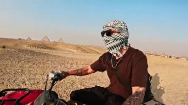 Genç yetişkinler ikonik giza piramitlerinin yakınındaki kumlu arazide geziniyorlar. Geniş mavi gökyüzü altında dörtlü bisiklet sürüyorlar. Maceracı bir turizm anı yaşıyorlar.