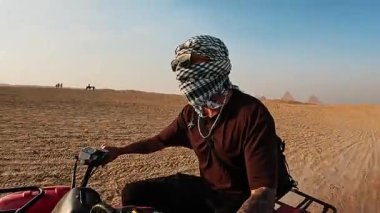 Dörtlü bisikletteki engebeli çöl arazisinde gezinen turistler, parlak mavi gökyüzü altındaki ikonik giza piramitlerini geçerek, heyecan verici Mısır maceralarını ve kültürel manzaraları tecrübe ediyorlar.