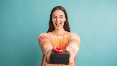 Renkli süveterli, gülümseyen bir kadın kırmızı kurdeleli küçük bir hediye kutusu sunuyor.