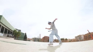 Breakdansçı dinamik akrobatik hareketler yapıyor, şehir planına karşı mimari unsurlarla yüksek enerjili sokak dansı becerileri sergiliyor.