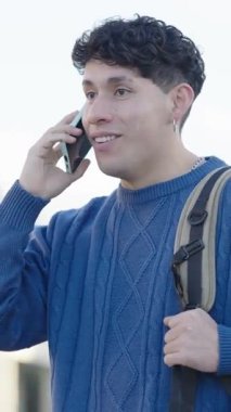 Mavi kazaklı genç bir erkek şehir caddesinde yürüyor. Sırt çantası, küpe ve kıvırcık saçlarıyla akıllı telefonuyla konuşuyor. Dikey görüntüler