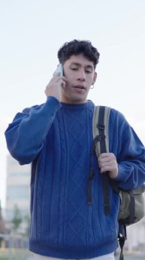 Üniversite öğrencisi sırt çantası taşıyor ve dışarıda yürürken akıllı telefonuyla konuşuyor. Dikey görüntüler