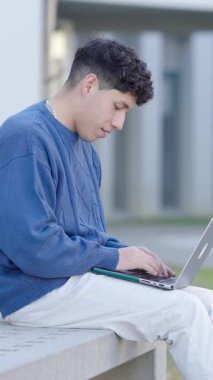 Genç erkek öğrenci kampüs bankında otururken dizüstü bilgisayarda çalışıyor, gündüz yoğunlaşarak akademik proje üzerinde çalışıyor. Dikey görüntüler