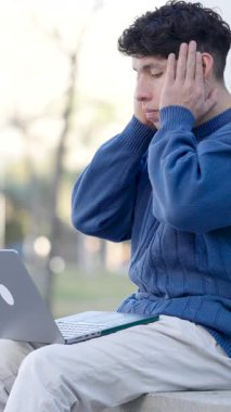 Yeşil park ortamında dizüstü bilgisayar çalışmalarıyla boğuşan genç erkek öğrenci, akademik stres ve dijital iş sorunları gösteriyor. Dikey görüntüler