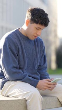 Genç Latin erkek bankta oturuyor, açık hava ortamında akıllı telefonla uğraşıyor, dijital iletişim ve eğlence için mobil cihaz kullanıyor. Dikey görüntüler