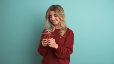 Sarı saçlı, kestane rengi kazaklı, gülümseyen genç bir kadının portresi mavi arka planda dikilirken cep telefonundan mesajlar okuyor.