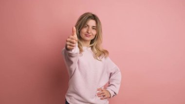 Yavaş çekimde, pembe arka planda, genç, kendine güvenen bir sarışının başparmağıyla el kol hareketi yaptığı bir video.