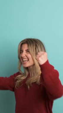Kestane rengi kazaklı heyecanlı genç bir kadın. Sarışın, yumruklarını sıkarak başarısını kutlarken mavi arka planda duran kameraya bakıyor.