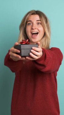 Kestane rengi kazaklı, uzun sarı saçlı, gülümseyen genç bir kadının portresi mavi arka planda sürpriz bir hediye kutusu alıyor.