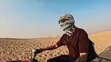 Genç erkek turist, kumlu giza piramitlerini dört tekerli bisikletle geçiyor, parlak mavi gökyüzü altında maceraperest çöl keşifleri yaşıyor.