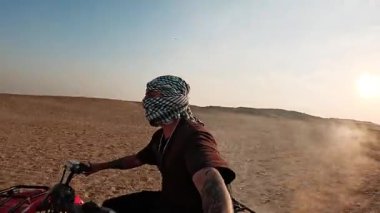 Geleneksel keffiyeh giyen genç turist dörtlü bisiklete biniyor, toz bulutları üretiyor ve gün batımında altın ışıklı çöl manzarasını aşıyor.