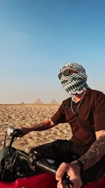 Antik giza piramitleri yakınlarındaki kumlu arazide gezen maceraperest bir gezgin geleneksel keffiyeh giyer ve güneş gözlüklü koruyucu gözler takar ve güneşli çöl manzarasında dört tekerli bisiklet sürer.