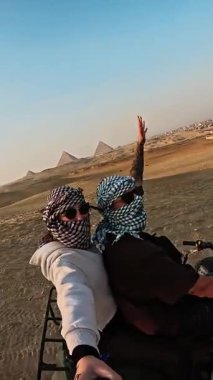 Maceraperest turistler ikonik giza piramit kompleksini çevreleyen kumlu arazide dörtlü bisiklete binerek parlak güneş ışığı altında tarihi Mısır manzarasını deneyimlerler.