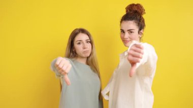 Onaylamadığını belirten iki genç kadın canlı sarı bir arkaplan karşısında el kol hareketi yapıyor.