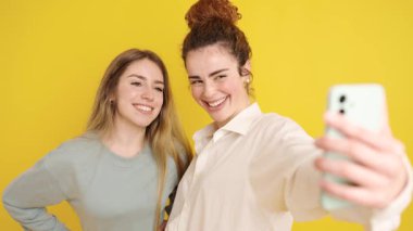 İki neşeli genç kadın bir dizi selfie çekiyor, farklı pozlar ve ifadeler deniyor, parlak sarı bir arka planda izole edilmiş.