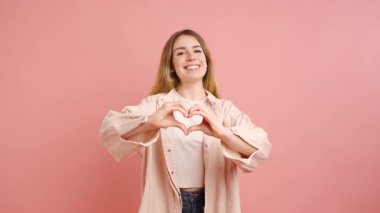 Sevgisini ve sevgisini ifade eden genç bir kadın, elleriyle kalp şeklinde jestler yapar ve canlı pembe bir arka plana öpücük atar.