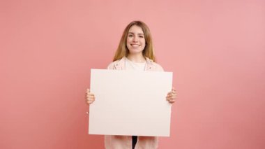 Genç bir kadın canlı pembe stüdyo arka planına karşı beyaz bir tabela tutuyor, gülümsüyor ve reklam veya reklam için boş bir alan sunuyor.