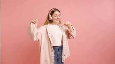 Kulaklıkla müzik dinleyen, pembe arka planda dans eden ve gülümseyen mutlu genç bir kadın.