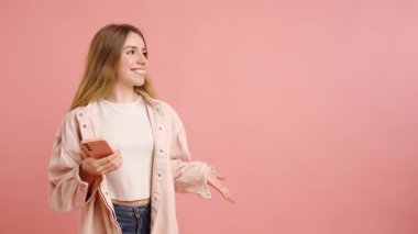 Akıllı telefonu tutan genç bir kadın konuşurken ve hareketli bir şekilde pembe stüdyo geçmişine bakarak neşeli bir enerjiyle kendini ifade ediyor.