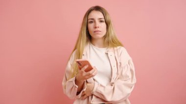 Uzun sarı saçlı, gündelik kıyafetler giyen genç bir kadın, pembe arka planda akıllı telefonunu kullanırken çeşitli duygular gösteriyor.