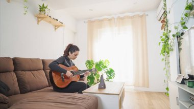 Evde klasik gitar pratiği yapan müzisyen, rahat bir oturma odasındaki kanepede dinlenirken bir tablette online ders veriyor.