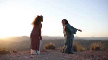 İki kadın gün batımında güzel bir çöl manzarasında dans edip zıplıyorlar.
