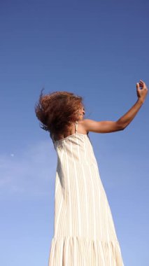 Kendine güveni tam genç bir kadın açık mavi gökyüzüne karşı özgürlük ve neşe içinde dans eder, kaygısız ve hayat dolu bir ruhu somutlaştırır.