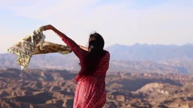 Kadın sanatçı zarif bir şekilde şalla dans ediyor, canlı kırmızı elbiseler giyiyor, geniş çöl arazisinin ortasında dramatik dağ zeminiyle, özgürlük ve iç ahengi ifade ediyor.