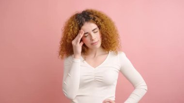 Kızıl saçlı kadın baş ağrısı çekiyor. Şakaklarına ve alnına dokunuyor. Pembe arka planda elleri var.