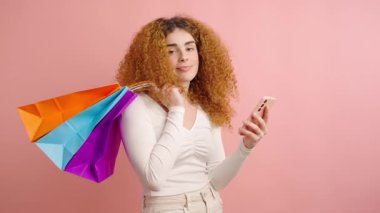 Kıvırcık saçlı, renkli alışveriş poşetleri taşıyan ve pembe arka planda dururken akıllı telefon kullanan neşeli, genç bir kadın.