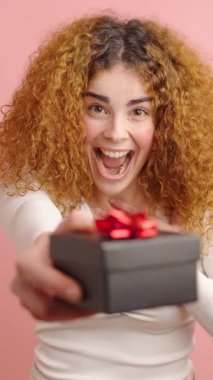 Kıvırcık saçlı kadın, elinde pembe bir arka planda izole edilmiş bir hediye kutusu tutarken mutluluk ifadeleri sergiliyor.