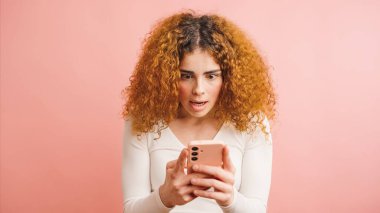 Şaşırmış, kıvırcık saçlı, ağzı açık duran ve pembe arka planda mesaj atan cep telefonu ekranına bakan İspanyol bir kadın.