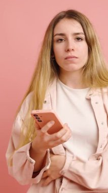 Genç bir kadın, akıllı telefonuyla pembe arka planda etkileşime girerken çeşitli olumsuz duygular gösterir.