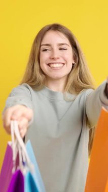 Neşeli genç bir kadın canlı alışveriş torbaları sergiliyor, mutluluğu yayıyor ve son zamanlarda parlak sarı arka planda yapılan alışverişleri sergiliyor.