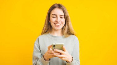 Mutlu genç kadın, canlı sarı bir arka planda dururken akıllı telefonunu kullanıyor ve gülümsüyor.