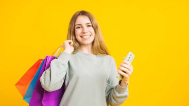 Sarı arka planda izole edilmiş bir mağazadan satın aldıktan sonra mutlu bir müşteri cep telefonu kullanıyor ve renkli alışveriş çantaları taşıyor.