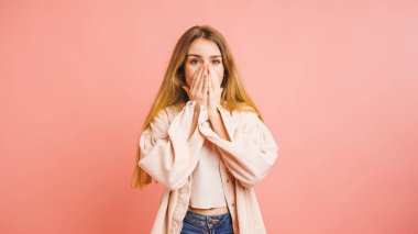 Şaşırmış genç bir kadının elleriyle ağzını kapamasının stüdyo portresi, canlı pembe bir arka planda şok ve güvensizlik ifadesidir.