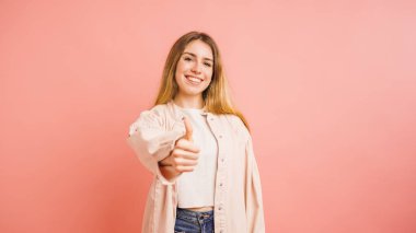 Mutlu genç bir kadın işaret ediyor onay ve başarı işareti, pembe arka planda gülümsüyor.