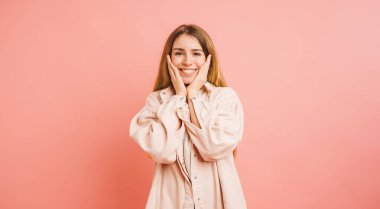 Elleriyle yanaklarını tutarak gülümseyen ve pembe bir arka planda mutluluğu ifade eden neşeli genç bir kadının stüdyo portresi.