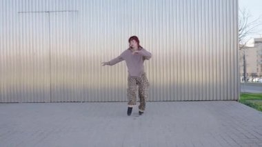 Metal şehir duvarına karşı modern koreografi sergileyen kızıl saçlı dansçı ritmik enerji ve tutkuyla yaratıcı hareketi ifade ediyor.