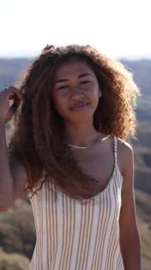 Uzun kıvırcık saçları olan genç bir kadın, neşeli bir şekilde iplikleri savurarak güneşli açık hava manzarasında duruyor, tasasız bir mutluluk yayıyor.