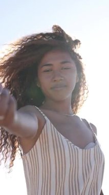 Rüzgarın süpürdüğü yaz elbisesi kıvırcık saçlı kadın özgürce dans ediyor, parlak mavi gökyüzü altında saf neşe ve gençlik enerjisi yayıyor.