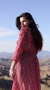 Kumral manken kırmızı çiçekli elbise giyiyor. Manzaralı çöl manzarasında duruyor. Rüzgâr, kurak vadi arkaplanına doğru uzun saçlar savuruyor.